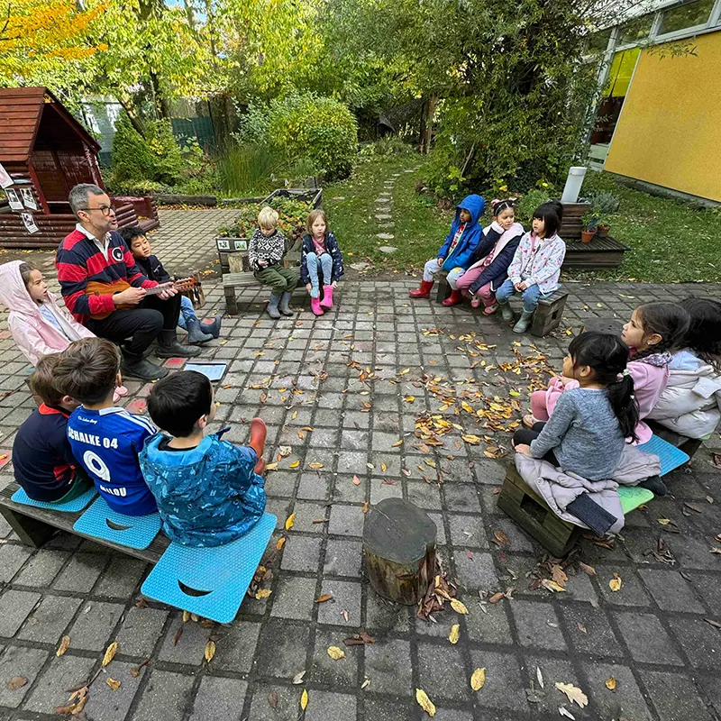 An educator plays a stringed instrument for a circle of young kindergarten students outside ISD campus.